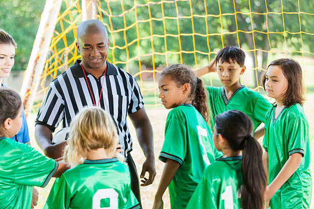 Mid adult African American youth soccer referee explains the rules of the game to the team dressed in green jerseys. Their female coach listens as well. The team is a coed team. They are standing behind the soccer goal.